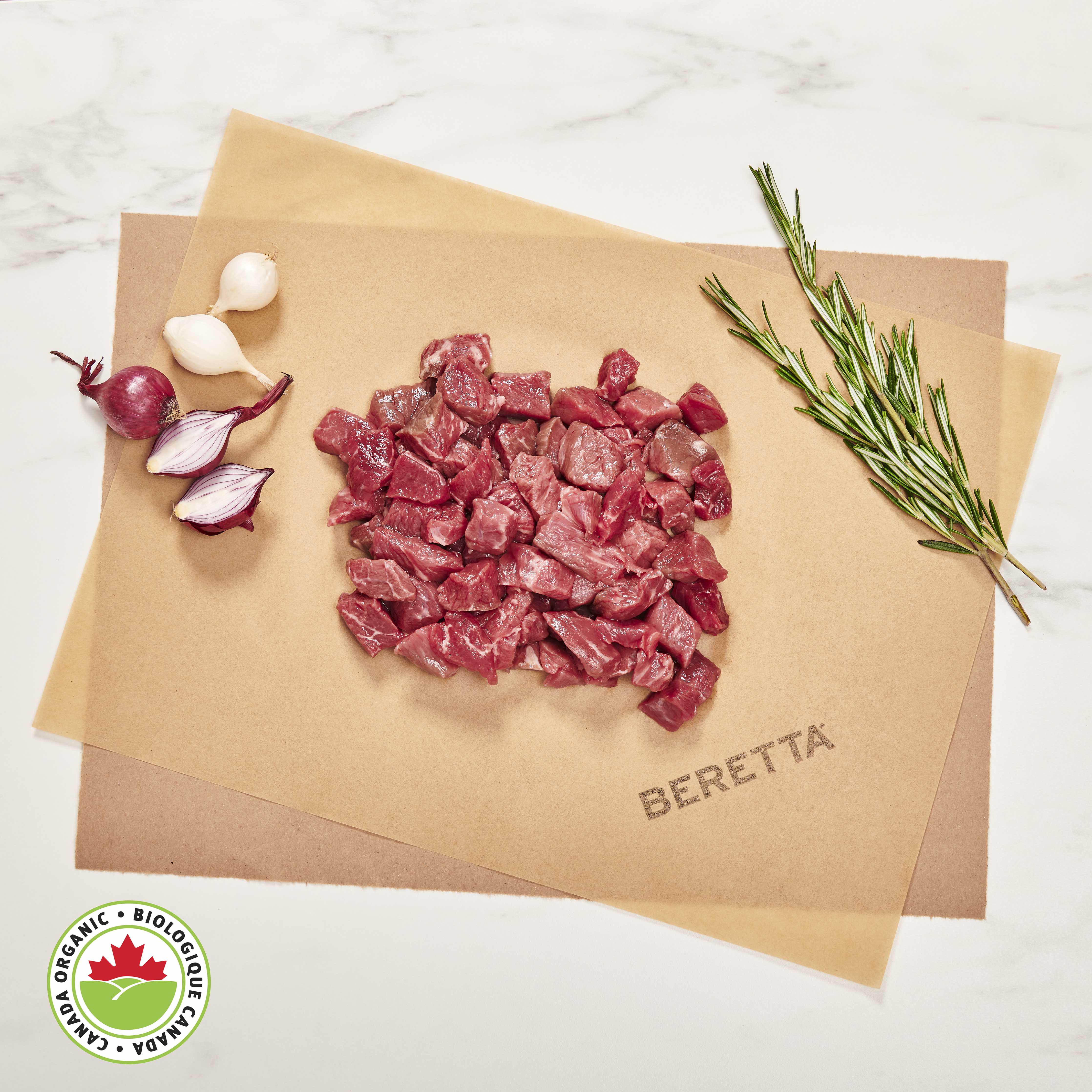 Chopped pieces of lean stewing beef on a paper, surrounded by garlic cloves and a sprig of rosemary, with the brand 'BERETTA' visible on the paper.