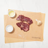 Raw beef bavette steaks on a wooden cutting board with Montreal spice, accompanied by a small white bowl with a liquid, next to a bulb of garlic and a piece of onion, on a marble surface.