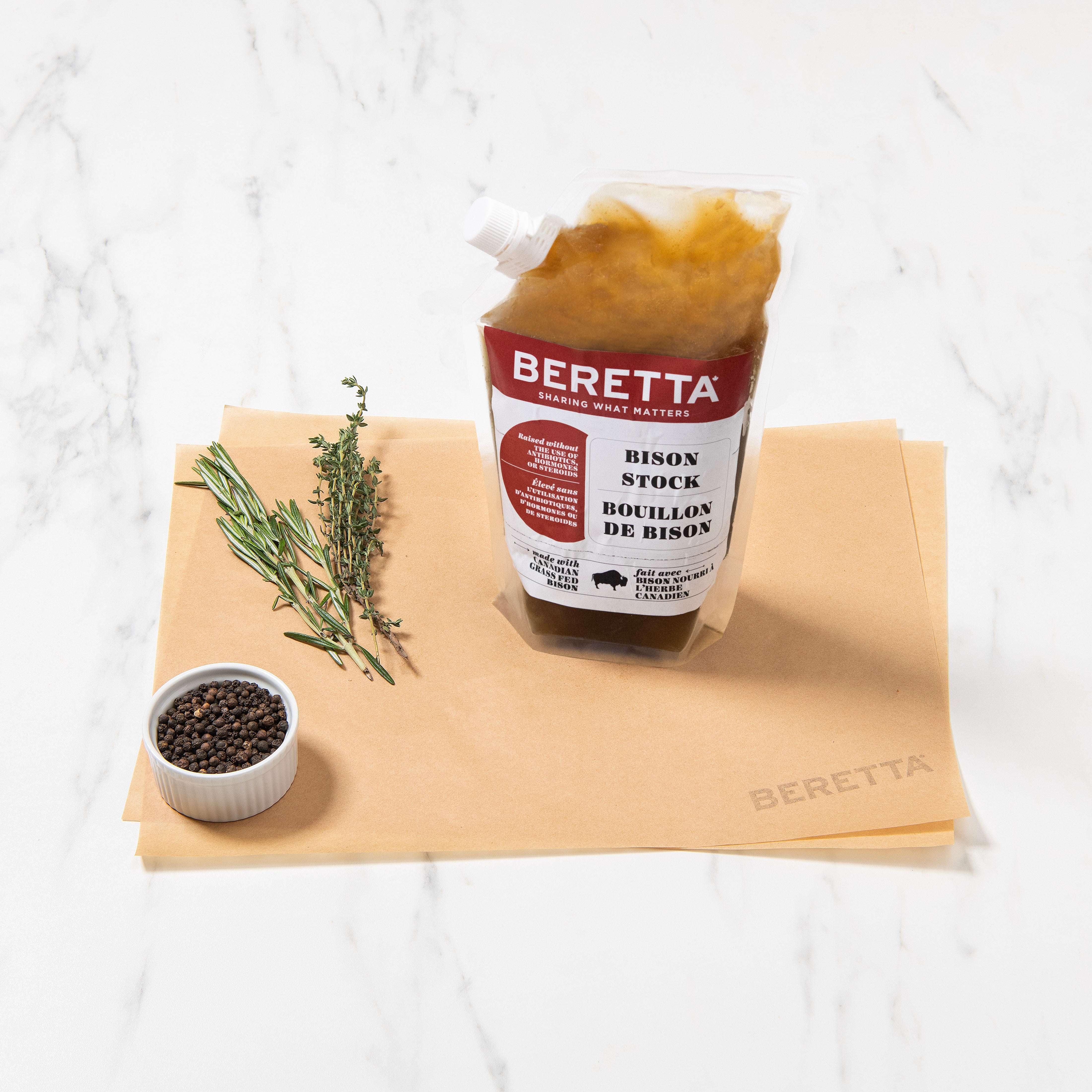 A clear package of Beretta bison stock, with a label, on a marble surface, alongside herbs and spices, and a small white bowl of seasoning.