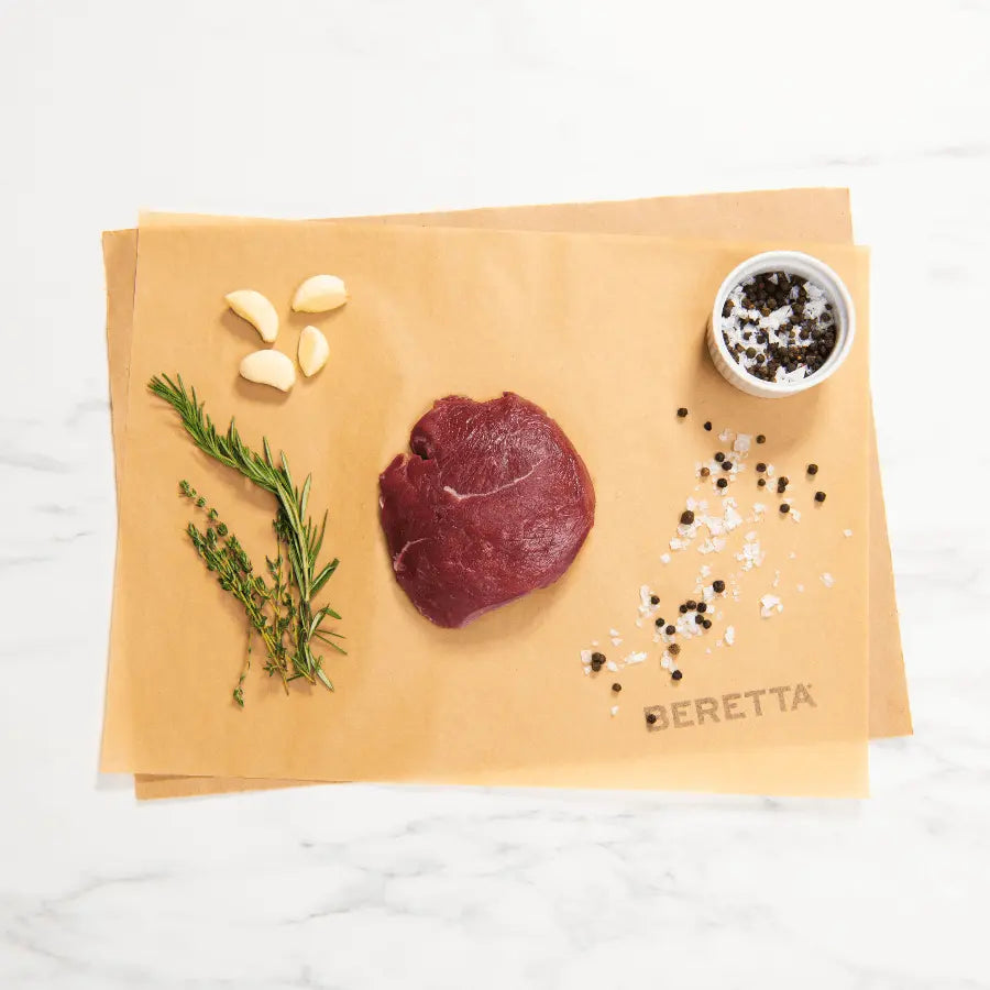 A raw bison top sirloin steak on brown paper, surrounded by seasonings, herbs, and garlic cloves, with the brand name 'BERETTA' visible on the paper.