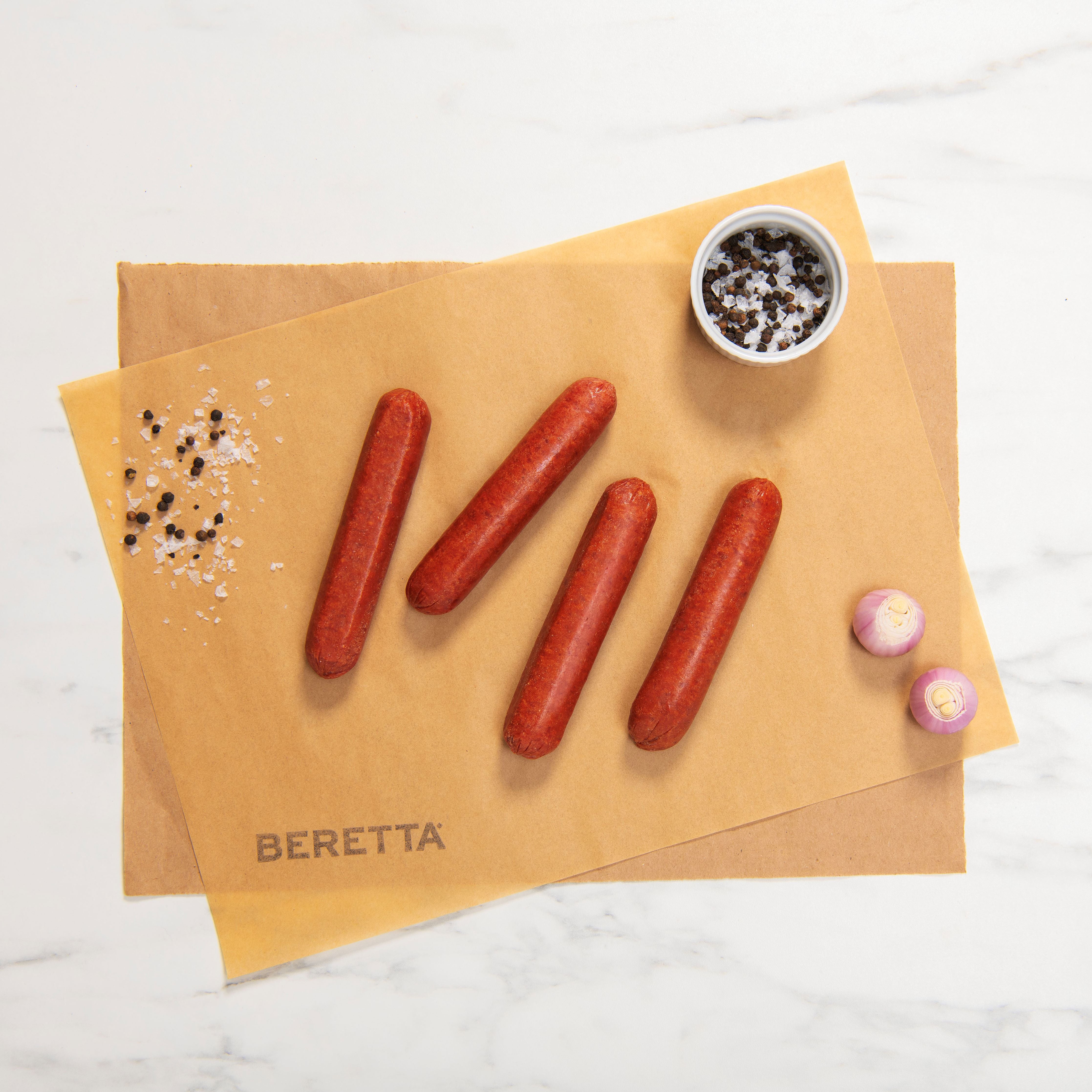 A pack of four jumbo bison hot dogs on a paper-like surface with seasonings on the side, branded with 'BERETTA' at the bottom.
