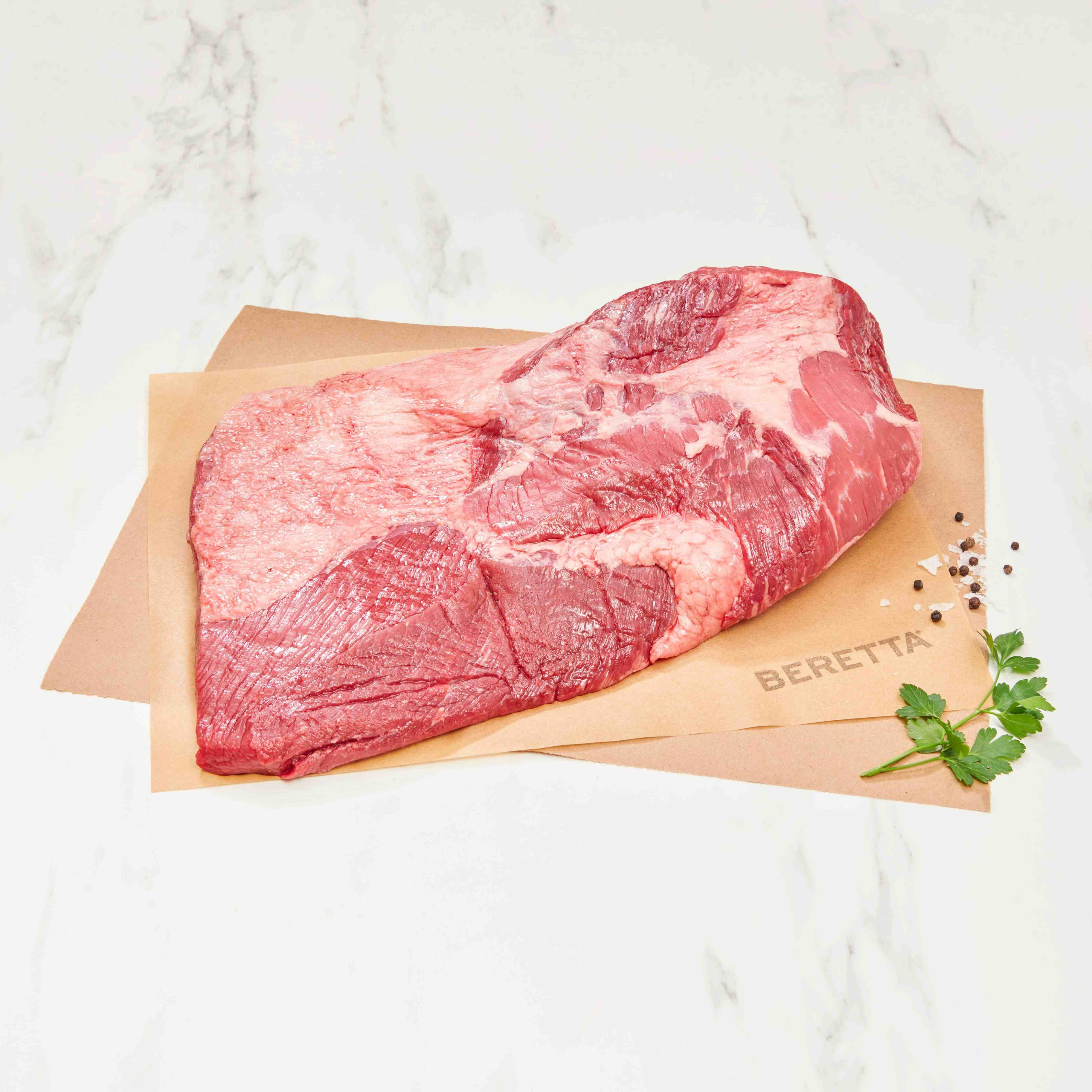 A raw beef brisket cut with visible marbling, placed on tan butcher paper branded BERETTA, on a white marble surface with a sprig of parsley and peppercorns nearby.