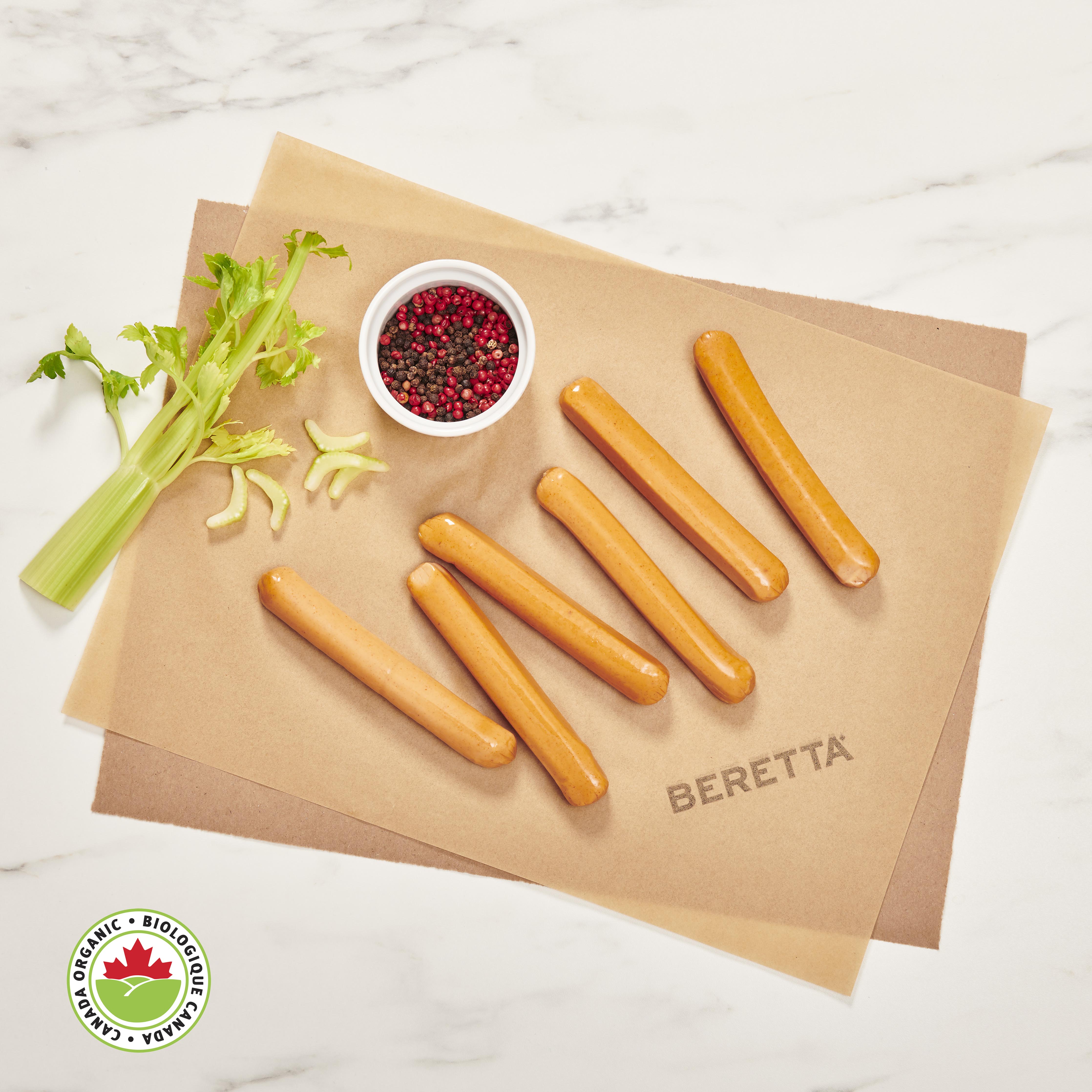 A pack of six chicken hot dogs on a beige paper against a white marble background, accompanied by celery sticks and a small bowl of spices. The brand 'BERETTA' is visible on the paper.